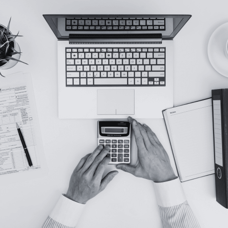 Vista superior de una mesa de escritorio con las manos de hombre sobre una calculadora frente a un ordenador portátil simulando el cálculo de impuestos y fiscalidad de una startup